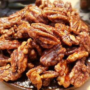 Puede incluir: Primer plano de un plato lleno de nueces pecanas caramelizadas. Las nueces son de un color marr&oacute;n intenso, cubiertas con un glaseado brillante y caramelizado. El plato es de color marr&oacute;n oscuro y tiene un aspecto ligeramente r&uacute;stico. La imagen est&aacute; bien iluminada, resaltando la textura y los detalles de las nueces.