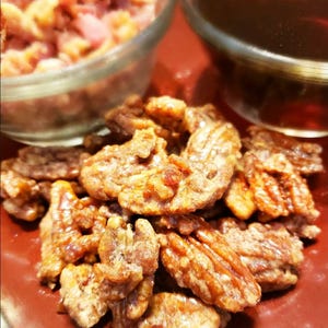 Puede incluir: Primer plano de nueces pecanas confitadas, con un glaseado brillante de color marr&oacute;n dorado, dispuestas sobre un plato rojo. Dos cuencos de vidrio en el fondo, uno con nueces picadas y el otro con un l&iacute;quido oscuro.