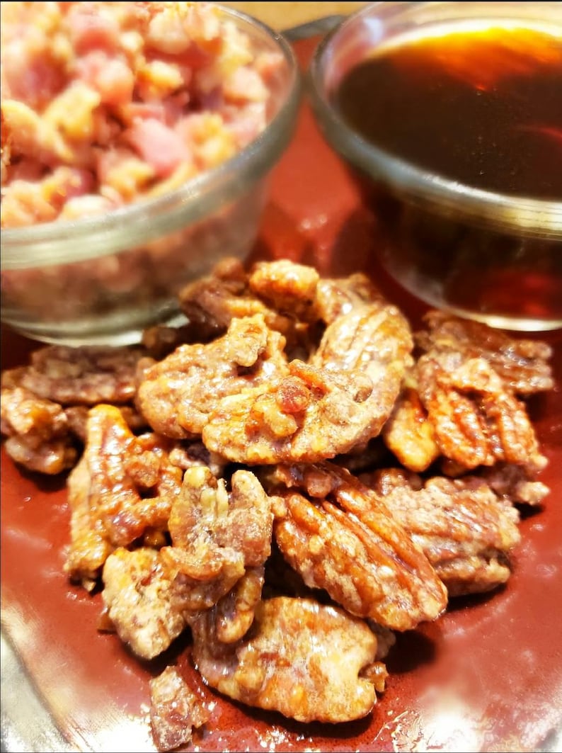 Puede incluir: Primer plano de nueces pecanas confitadas, con un glaseado brillante de color marr&oacute;n dorado, dispuestas sobre un plato rojo. Dos cuencos de vidrio en el fondo, uno con nueces picadas y el otro con un l&iacute;quido oscuro.