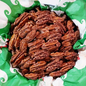 K&ouml;nnte beinhalten: Nahaufnahme eines Beh&auml;lters gef&uuml;llt mit kandierten Pekann&uuml;ssen. Die Pekann&uuml;sse haben eine satte braune Farbe und scheinen mit einer zuckerhaltigen Glasur &uuml;berzogen zu sein. Der Beh&auml;lter ist mit gr&uuml;nem und wei&szlig;em Dekopapier ausgekleidet.