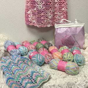May include: A collection of colorful yarn skeins with pink labels, a crocheted garment, and a clear bag with a pink insert. The yarn is in various colors, including blue, green, pink, and yellow. A crocheted garment in pink and white hangs in the background.