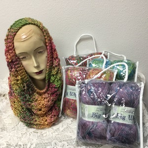 May include: A colorful crocheted cowl in shades of green, pink, and orange. The cowl is displayed on a mannequin head.  There are four bags of yarn in the background, two of which are labeled "Big  Wool".