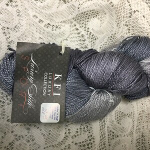 May include: A skein of KFI Luxury Collection "Spoo" yarn in a silver and gray ombre colorway. The label reads "KFI Luxury Collection Spoo 225 yds."