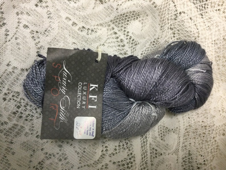May include: A skein of KFI Luxury Collection "Spoo" yarn in a silver and gray ombre colorway. The label reads "KFI Luxury Collection Spoo 225 yds."