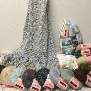 May include: A blue and beige knitted scarf with fringe, along with a bag of yarn in various colors, including blue, beige, brown, and green. The yarn is labeled "Louisa Harding".