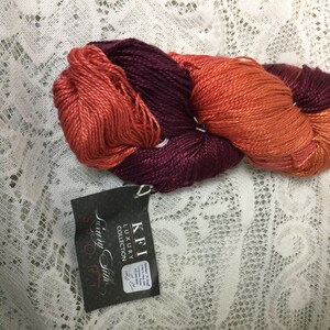 May include: A skein of yarn in two colors, a deep burgundy and a burnt orange. The yarn is labeled "KFI Luxury Collection" and "Luxury Silk".