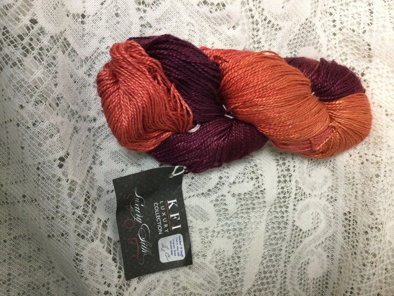 May include: A skein of yarn in two colors, a deep burgundy and a burnt orange. The yarn is labeled "KFI Luxury Collection" and "Luxury Silk".