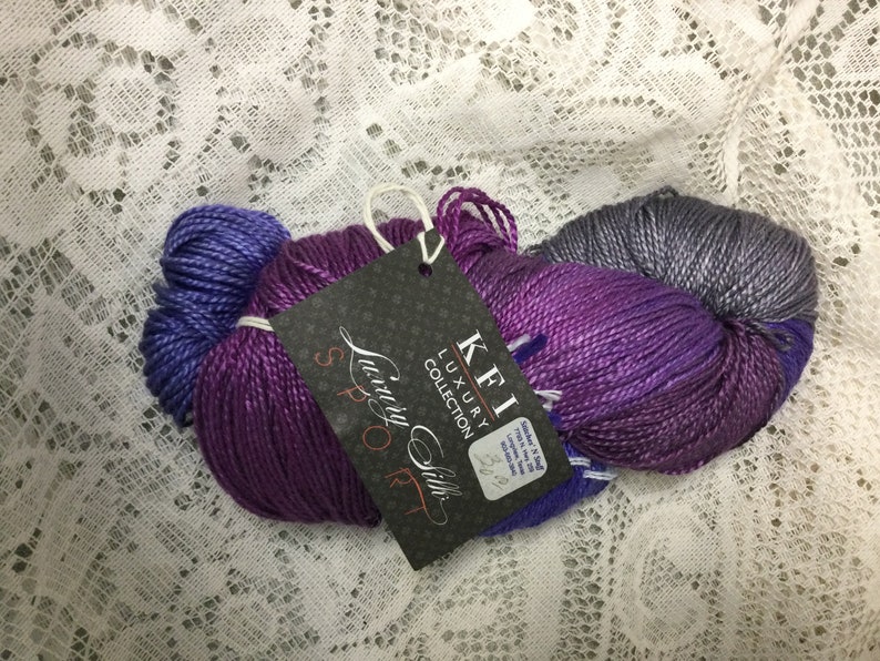 May include: A skein of purple, blue, and gray yarn with a label that reads "KFI Luxury Collection Luxury Silk Sport 32/2".