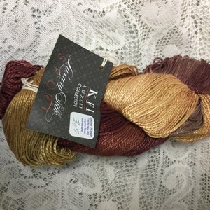 May include: A skein of KFI Luxury Collection Sport Silk yarn in shades of brown, gold, and burgundy. The label reads "KFI Luxury Collection Sport Silk 70% Silk & 30% Cashmere 1.5 oz (42.5g) 220 yds (201m)"
