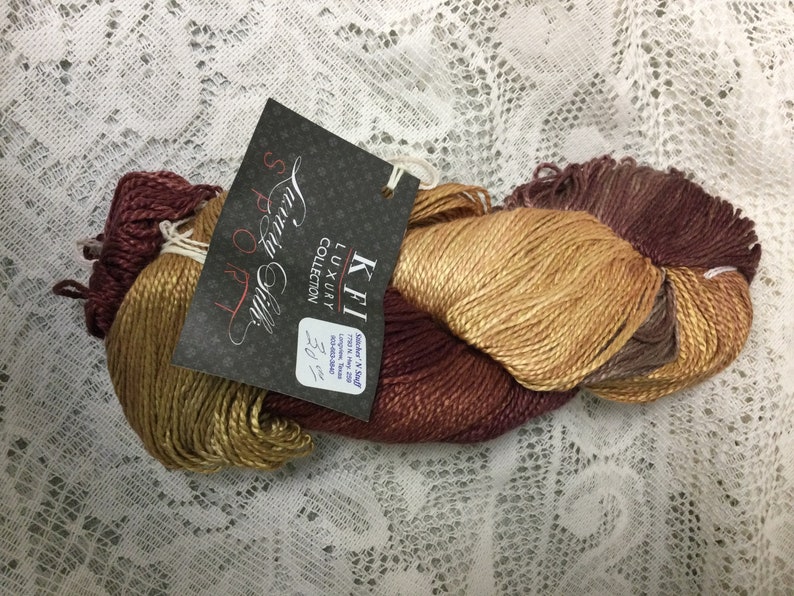 May include: A skein of KFI Luxury Collection Sport Silk yarn in shades of brown, gold, and burgundy. The label reads "KFI Luxury Collection Sport Silk 70% Silk & 30% Cashmere 1.5 oz (42.5g) 220 yds (201m)"