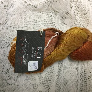 May include: A skein of KFI Luxury Collection "Sunday Silk" yarn in the "Spook" colorway. The yarn is a blend of orange, brown, and gold shades.