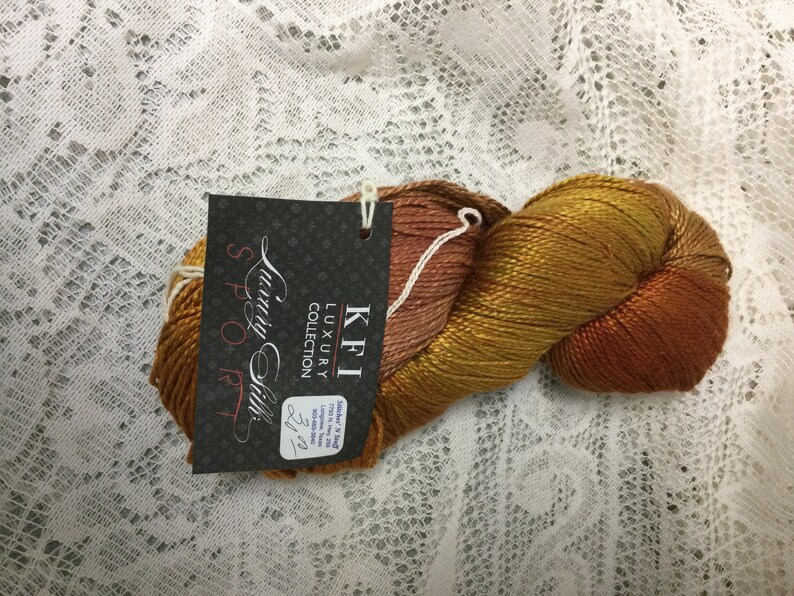 May include: A skein of KFI Luxury Collection "Sunday Silk" yarn in the "Spook" colorway. The yarn is a blend of orange, brown, and gold shades.