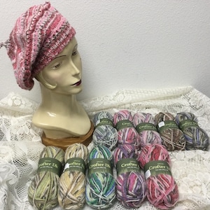 May include: A pink, gray, and white knit beret sits on a mannequin head. Several skeins of colorful yarn are arranged in front of the mannequin. The yarn colors include green, blue, pink, and brown. The yarn is labeled "Crofter DK".