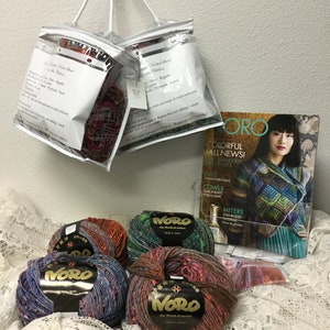 May include: Four skeins of Noro yarn in various colors, including red, blue, green, and brown. The yarn is arranged on a white lace tablecloth. The yarn is labeled "Noro, the world of nature, made in Japan". There is a magazine in the background with the title "Noro, Colorful Fall News! Standout Styles".