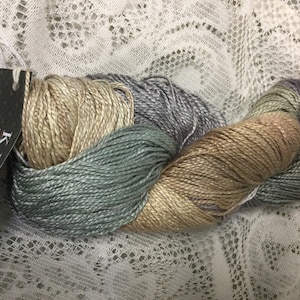 May include: A skein of K.H.I. Luxury Collection silk yarn in a multi-colored ombre pattern of beige, light blue, and lavender.