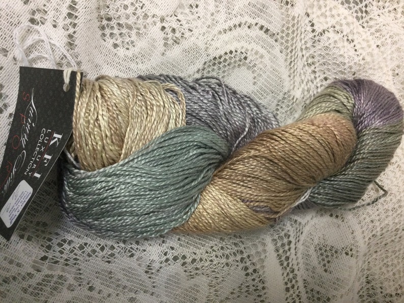May include: A skein of K.H.I. Luxury Collection silk yarn in a multi-colored ombre pattern of beige, light blue, and lavender.