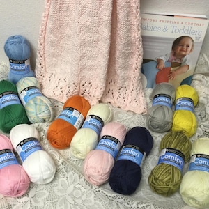 May include: A collection of colorful yarn skeins in various shades, including pink, green, orange, and blue, alongside a pink knitted baby blanket and a book titled "Comfort Knitting & Crochet Babies & Toddlers."