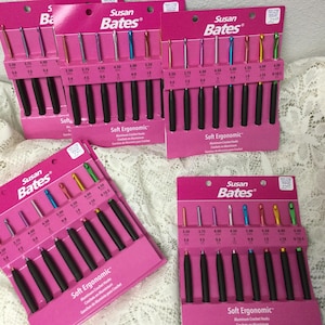 May include: Multiple sets of Susan Bates Soft Ergonomic crochet hooks are displayed. Each set, presented on a pink card, includes hooks in various sizes with black handles and colorful tips. The text "Soft Ergonomic" is visible.