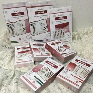 May include: Several boxes of Knit Pro Dreamz crochet and interchangeable needle sets. The boxes are white and burgundy with illustrations of the contents. The sets include needles, hooks, and accessories. The boxes are arranged on a white lace surface.