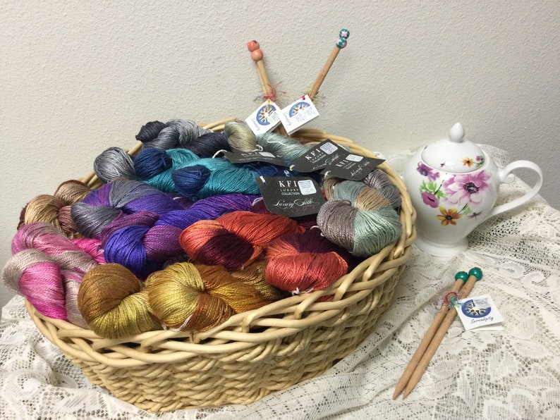 May include: A wicker basket filled with colorful skeins of yarn, some with metallic threads. The yarn is labeled "KFI Luxury Collection" and "Simply Silk". There are also two pairs of knitting needles with beads on the ends.