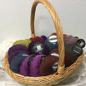 May include: A wicker basket filled with skeins of yarn in various colors, including purple, green, brown, and gray. The yarn is labeled "Classic Jo Sharp" and is arranged in a circular pattern.
