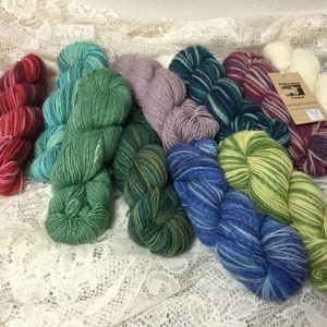 May include: Assortment of colorful yarn skeins in various shades of green, blue, purple, red, and white. The yarn is arranged in a semi-circle on a white lace tablecloth. The label on one skein reads "Moonshine Farm".
