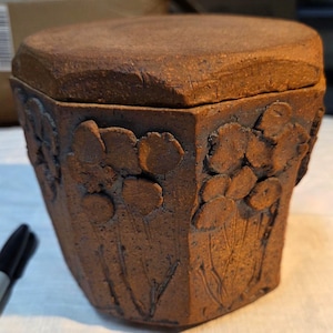 May include: A brown, hexagonal ceramic jar with a matching lid. The exterior features a textured, raised relief pattern of floral stems and rounded petals on each side. The clay has a matte, earthy finish with visible tool marks and rough edges.