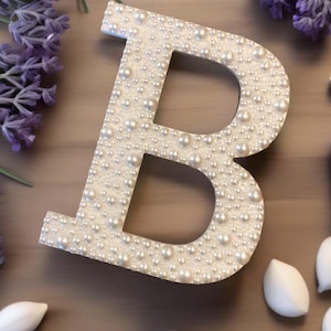 May include: A large, white letter "B" is covered in small, faux pearls. The letter is surrounded by purple lavender and white, leaf-shaped decorations, creating a decorative and elegant design. The letter is a perfect accent for home decor.