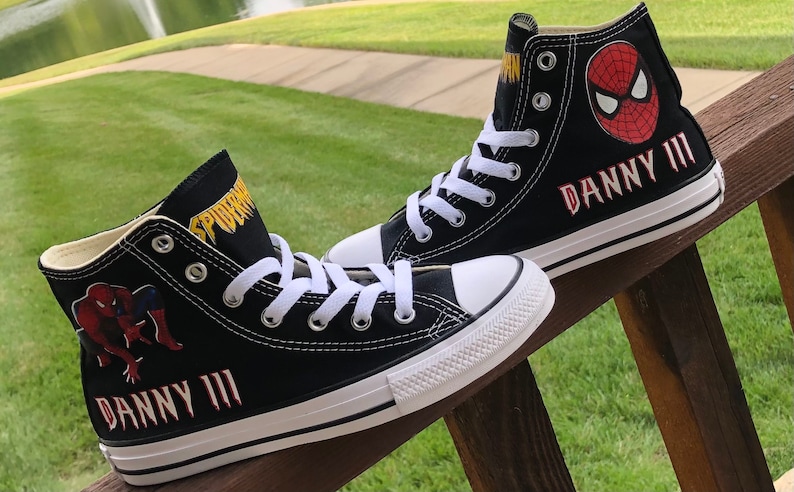 K&ouml;nnte beinhalten: Paar schwarze High-Top-Leinenschuhe mit Spider-Man-Grafiken. Die Schuhe haben wei&szlig;e Schn&uuml;rsenkel, eine wei&szlig;e Zehenkappe und den Namen "DANNY III" in Rot. Das Spider-Man-Logo und der Name befinden sich ebenfalls auf der Seite.