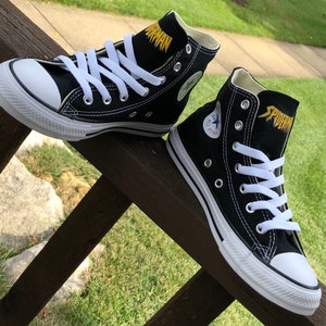 K&ouml;nnte beinhalten: Paar schwarze High-Top-Sneaker mit wei&szlig;en Schn&uuml;rsenkeln und Sohlen. Das Wort "SPIDERMAN" ist goldfarben auf der Zunge gestickt. Das ikonische All Star-Logo ist auf der Seite eines Schuhs zu sehen. Die Sneaker werden auf einer Holzoberfl&auml;che pr&auml;sentiert.