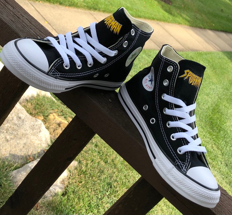 K&ouml;nnte beinhalten: Paar schwarze High-Top-Sneaker mit wei&szlig;en Schn&uuml;rsenkeln und Sohlen. Das Wort "SPIDERMAN" ist goldfarben auf der Zunge gestickt. Das ikonische All Star-Logo ist auf der Seite eines Schuhs zu sehen. Die Sneaker werden auf einer Holzoberfl&auml;che pr&auml;sentiert.