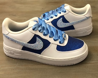 May include: White and blue sneakers with sparkling accents. The shoes feature a white base with blue glitter panels and a jeweled Nike swoosh. Light blue satin ribbon laces add a touch of elegance. The shoes have a white sole.