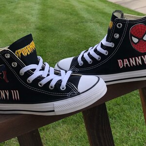 K&ouml;nnte beinhalten: Paar schwarze High-Top-Sneaker mit Spider-Man-Grafiken. Die Sneaker haben wei&szlig;e Schn&uuml;rsenkel, Sohlen und Zehenkappen. Der Name "DANNY III" ist rot auf die Seite jedes Schuhs gestickt. Das Wort "SPIDERMAN" ist gelb auf die Zunge gedruckt.