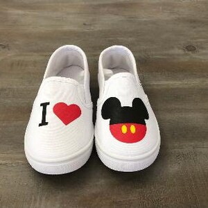 Puede incluir: Par de zapatos blancos sin cordones. Un zapato presenta el texto "I" con un coraz&oacute;n rojo. El otro zapato tiene un dise&ntilde;o de Mickey Mouse con orejas negras, una cara roja y botones amarillos. Los zapatos est&aacute;n sobre una superficie de madera.