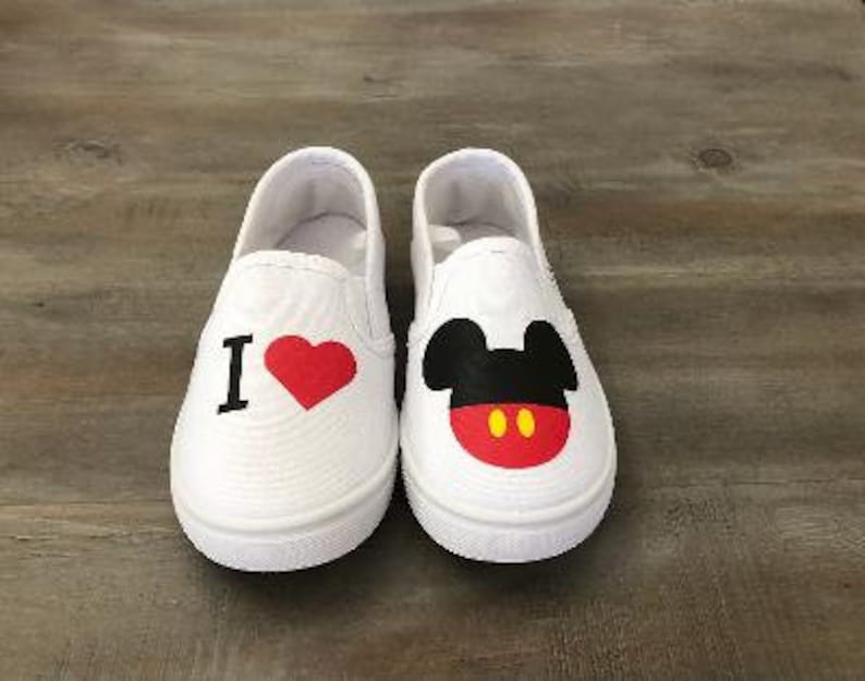 Puede incluir: Par de zapatos blancos sin cordones. Un zapato presenta el texto "I" con un coraz&oacute;n rojo. El otro zapato tiene un dise&ntilde;o de Mickey Mouse con orejas negras, una cara roja y botones amarillos. Los zapatos est&aacute;n sobre una superficie de madera.