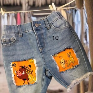 May include: Light blue denim shorts with frayed edges and orange patches featuring characters from The Lion King. The number "10" is printed on the shorts.