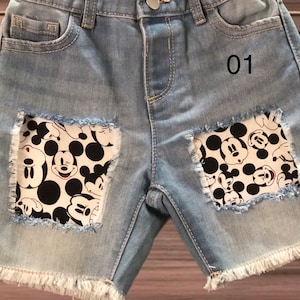 May include: Light blue denim shorts with frayed edges and two patches featuring a black and white Mickey Mouse pattern. The number "01" is printed on the right side of the shorts. These shorts are a fun and playful addition to any child's wardrobe.