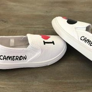 Puede incluir: Par de zapatos blancos de lona sin cordones. Un zapato tiene un coraz&oacute;n rojo y la letra "I" impresa. Ambos zapatos tienen el nombre "CAMERON" impreso en negro. Los zapatos est&aacute;n sobre una superficie de madera.