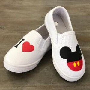Puede incluir: Zapatillas blancas sin cordones con dise&ntilde;o de Mickey Mouse. Un zapato tiene una "I" negra y un coraz&oacute;n rojo, el otro una cara de Mickey Mouse con orejas negras, boca roja y detalles amarillos. Sobre una superficie de madera.
