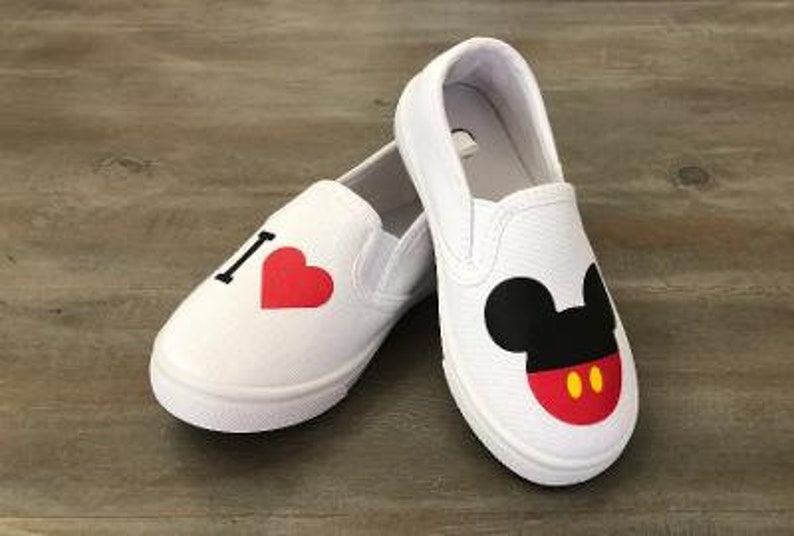 Puede incluir: Zapatillas blancas sin cordones con dise&ntilde;o de Mickey Mouse. Un zapato tiene una "I" negra y un coraz&oacute;n rojo, el otro una cara de Mickey Mouse con orejas negras, boca roja y detalles amarillos. Sobre una superficie de madera.