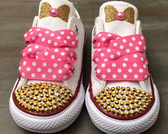 May include: A pair of white canvas sneakers with gold glitter bows and pink polka dot ribbon laces. The toe caps are embellished with gold rhinestones. A pink pearl is in the center of each bow.
