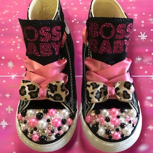 May include: A pair of black canvas sneakers with the words "BOSS BABY" in pink glitter. The shoes feature pink satin ribbon laces, leopard print bows, and are embellished with pink, white, and black pearls and rhinestones on the toes.