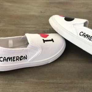 Puede incluir: Zapatillas blancas sin cordones con el texto "I ❤️ CAMERON" en negro y rojo. La palabra "CAMERON" tambi&eacute;n est&aacute; impresa en el lateral de cada zapatilla en negro. Las zapatillas est&aacute;n sobre una superficie de madera.