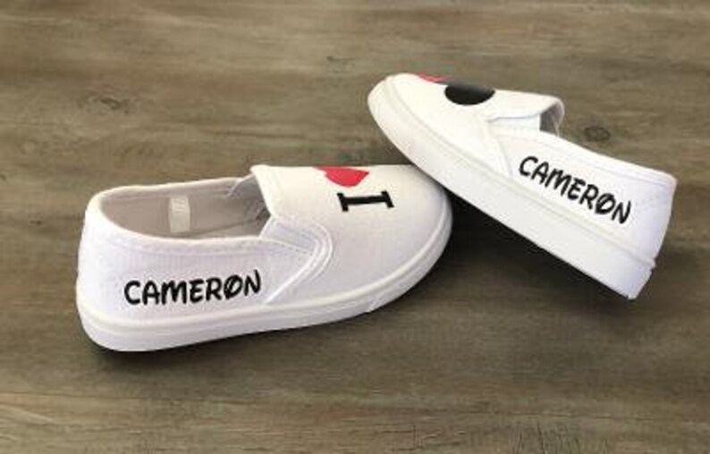 Puede incluir: Zapatillas blancas sin cordones con el texto "I ❤️ CAMERON" en negro y rojo. La palabra "CAMERON" tambi&eacute;n est&aacute; impresa en el lateral de cada zapatilla en negro. Las zapatillas est&aacute;n sobre una superficie de madera.