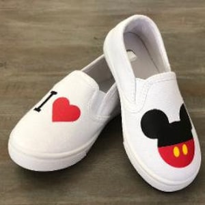 Puede incluir: Zapatillas blancas sin cordones con gr&aacute;ficos de dibujos animados. Un zapato tiene una "I" negra y un coraz&oacute;n rojo. El otro zapato presenta un dise&ntilde;o de Mickey Mouse con orejas negras, cara roja y botones amarillos. Sobre una superficie de madera.