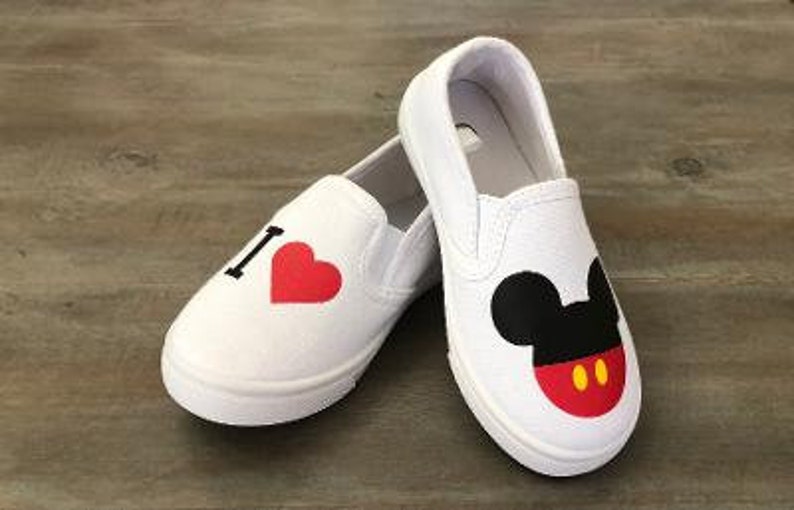 Puede incluir: Zapatillas blancas sin cordones con gr&aacute;ficos de dibujos animados. Un zapato tiene una "I" negra y un coraz&oacute;n rojo. El otro zapato presenta un dise&ntilde;o de Mickey Mouse con orejas negras, cara roja y botones amarillos. Sobre una superficie de madera.