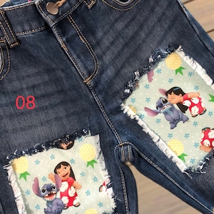 May include: Blue denim shorts with frayed edges and patches featuring the characters Lilo and Stitch. The patches have a light blue background with pineapples and floral patterns. The number "08" is embroidered in red.