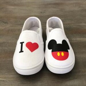 Puede incluir: Un par de zapatos blancos sin cordones. Un zapato presenta una "I" negra y un coraz&oacute;n rojo. El otro zapato muestra un dise&ntilde;o de Mickey Mouse con orejas negras, cara roja y detalles amarillos. Los zapatos est&aacute;n sobre una superficie de madera.