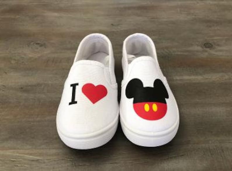 Puede incluir: Un par de zapatos blancos sin cordones. Un zapato presenta una "I" negra y un coraz&oacute;n rojo. El otro zapato muestra un dise&ntilde;o de Mickey Mouse con orejas negras, cara roja y detalles amarillos. Los zapatos est&aacute;n sobre una superficie de madera.