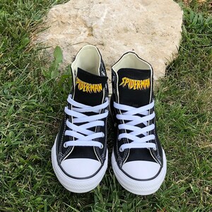 K&ouml;nnte beinhalten: Paar schwarze High-Top-Sneaker mit wei&szlig;en Schn&uuml;rsenkeln und Zehenkappen. Das Wort "SPIDERMAN" ist gelb auf der Zunge jedes Schuhs aufgedruckt. Die Schuhe stehen auf gr&uuml;nem Gras.